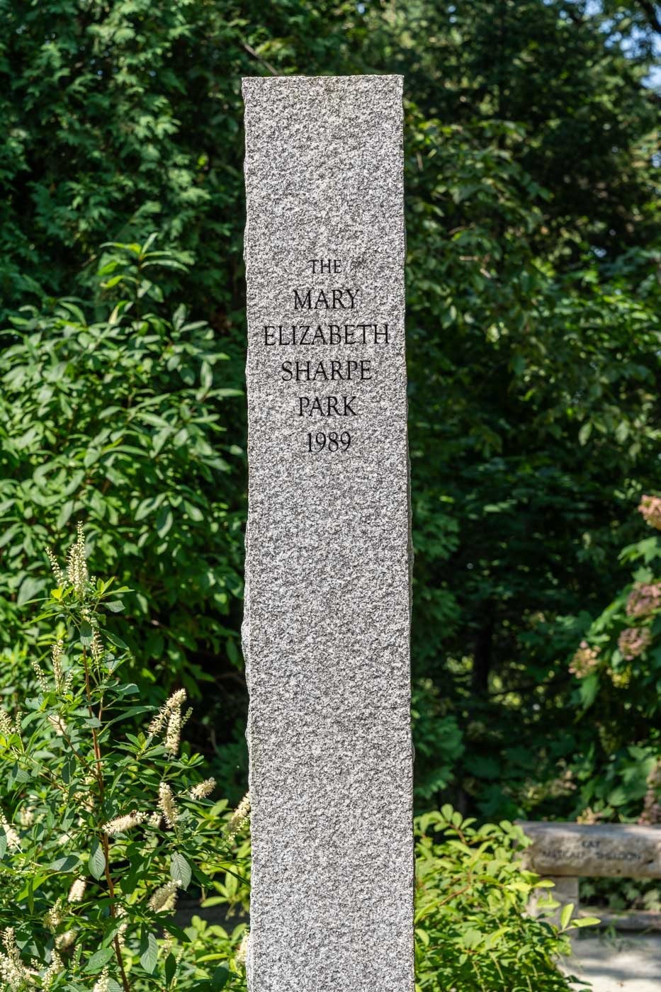 A tall, grey, rectangular memorial stone in the middle of a park.