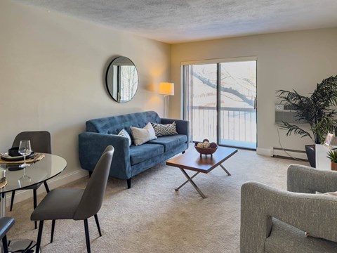 A living room with a blue couch, grey armchairs, a glass table, and a round mirror on the wall.