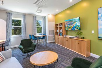 The Residences at Crosspoint cyber lounge in Lowell, MA includes a green accent wall with a mounted flat-screen TV, a wooden storage unit with open shelving, a dark green swivel chair, a round wood coffee table, and a high-top table with blue chairs by two curtained windows overlooking trees.