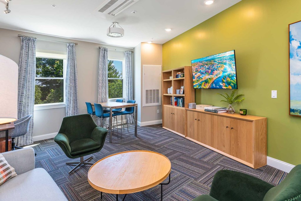 The Residences at Crosspoint cyber lounge in Lowell, MA includes a green accent wall with a mounted flat-screen TV, a wooden storage unit with open shelving, a dark green swivel chair, a round wood coffee table, and a high-top table with blue chairs by two curtained windows overlooking trees.