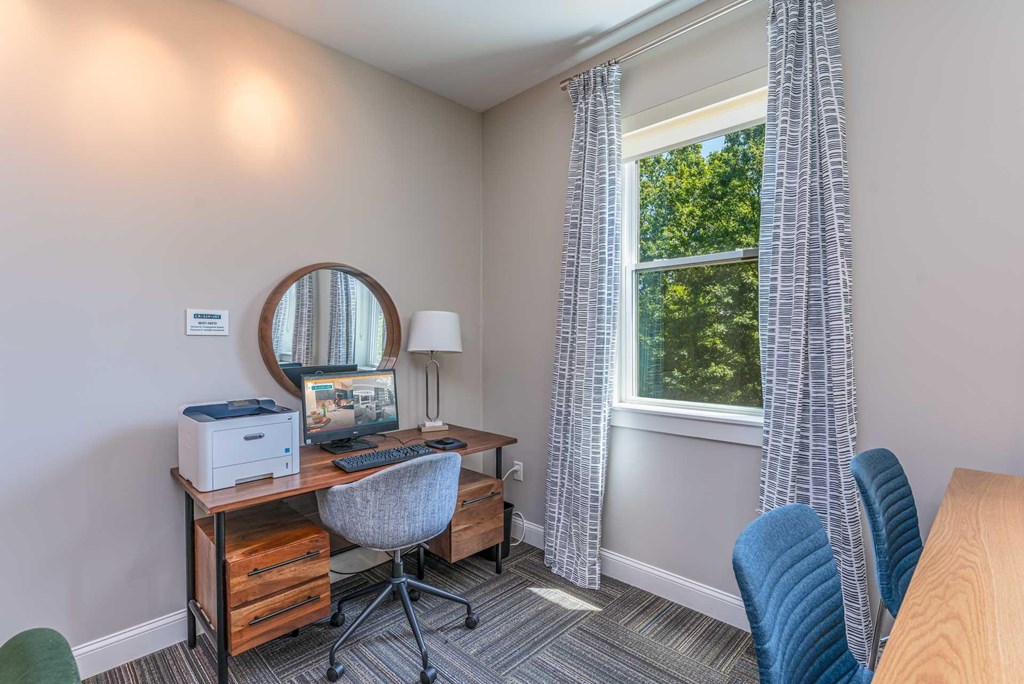 The Residences at Crosspoint study nook in the cyber lounge shows a wooden desk with a printer and desktop computer, a round wall mirror above, a gray upholstered swivel chair, and a floor lamp beside a curtained window looking out onto trees.