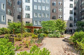 The Residences at Crosspoint central courtyard at 42 Wellman Street in Lowell, MA features paved walkways, raised garden beds, green shrubs, benches, and surrounding multi-story buildings clad in stone and metal siding.