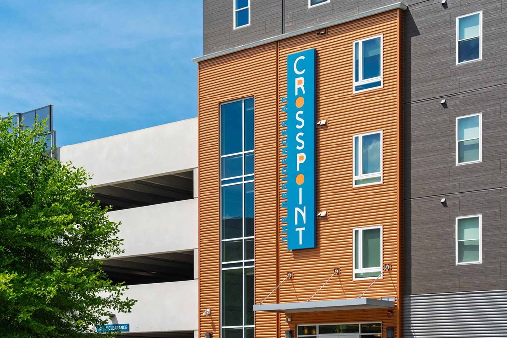 The Residences at Crosspoint apartment community at 42 Wellman Street in Lowell, MA features an orange corrugated-metal façade with a tall blue vertical sign reading "Crosspoint" mounted on the building next to a white concrete parking garage.
