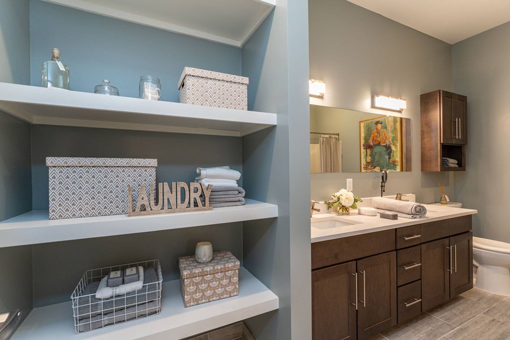 The Residences at Crosspoint laundry nook shows built-in white shelves holding decorative boxes, glass jars, and folded towels alongside a wooden "Laundry" sign, with a bathroom vanity visible in the background.