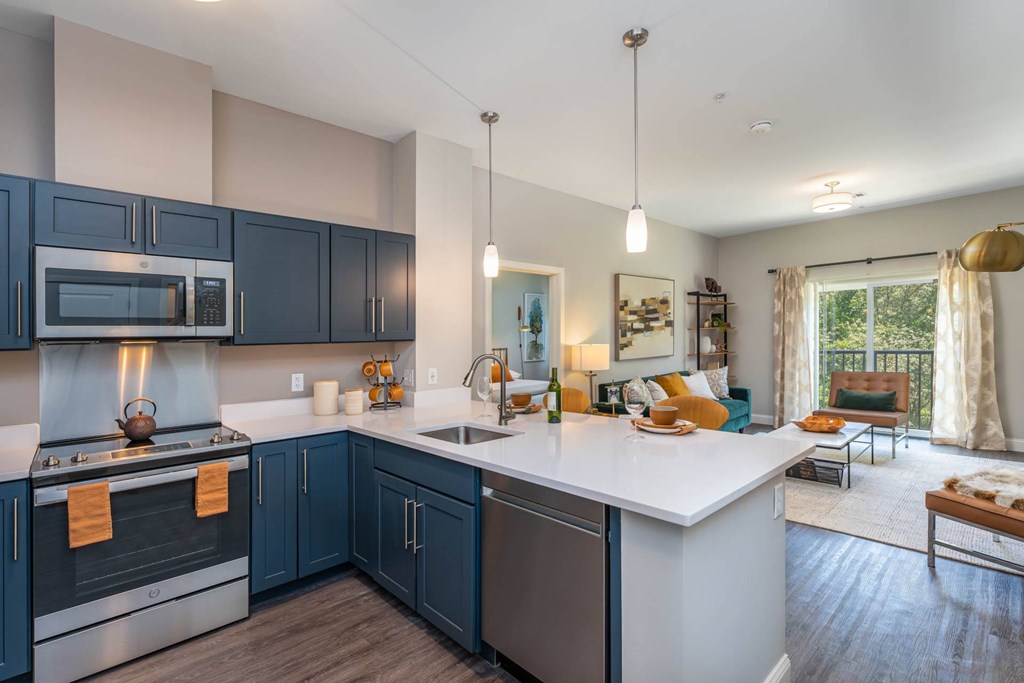 The Residences at Crosspoint open-plan kitchen and living area includes navy blue shaker cabinets, stainless steel appliances, a white island countertop with stools, and seating near a sliding glass door leading to a balcony.