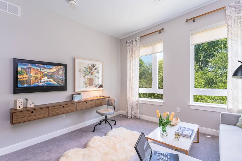 The Residences at Crosspoint living room corner features a wall-mounted flat-screen TV, abstract artwork above a floating wood console, a sheepskin rug on the floor, and a white coffee table holding a vase of yellow tulips.