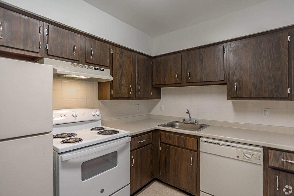 A kitchen with white appliances and wooden cabinets, disposals in each apartment, electric ranges, and dishwashers.