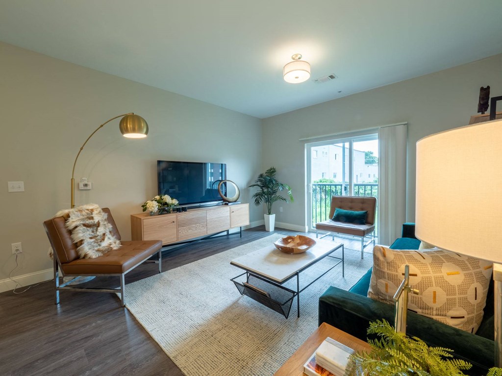 A bright living room with floor-to-ceiling sliding doors leading to a private balcony, furnished with modern seating and a flat-screen TV.