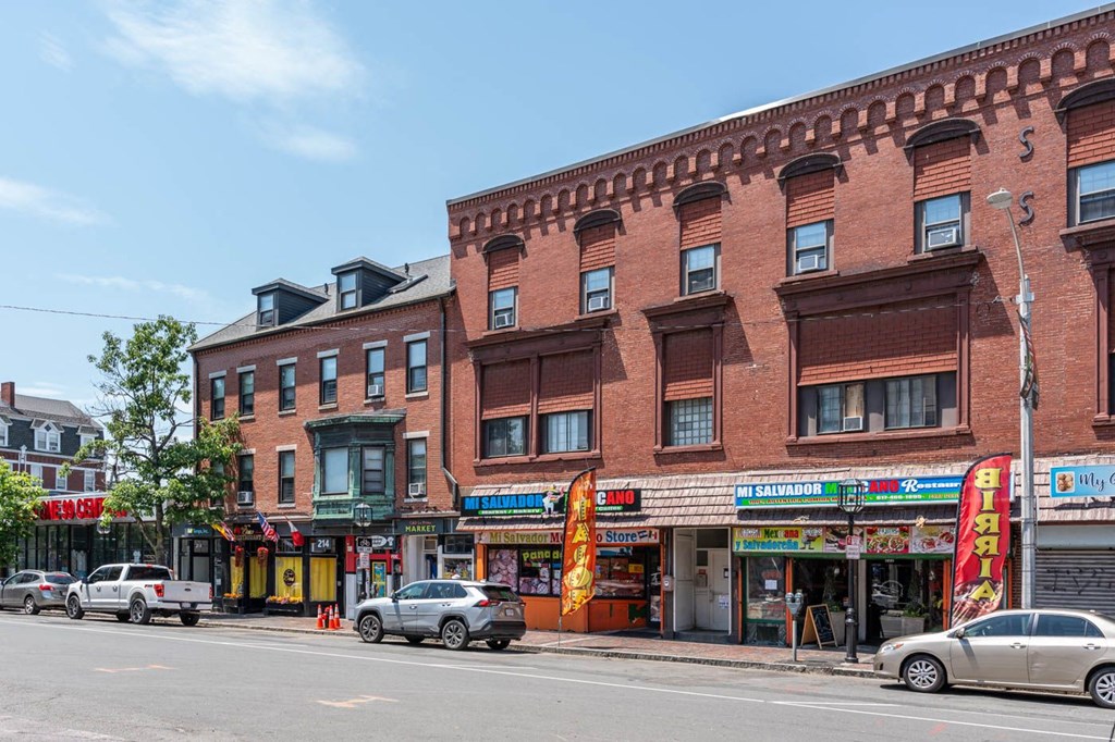 A street view of a city with cars parked and buildings with shops on the ground floor.