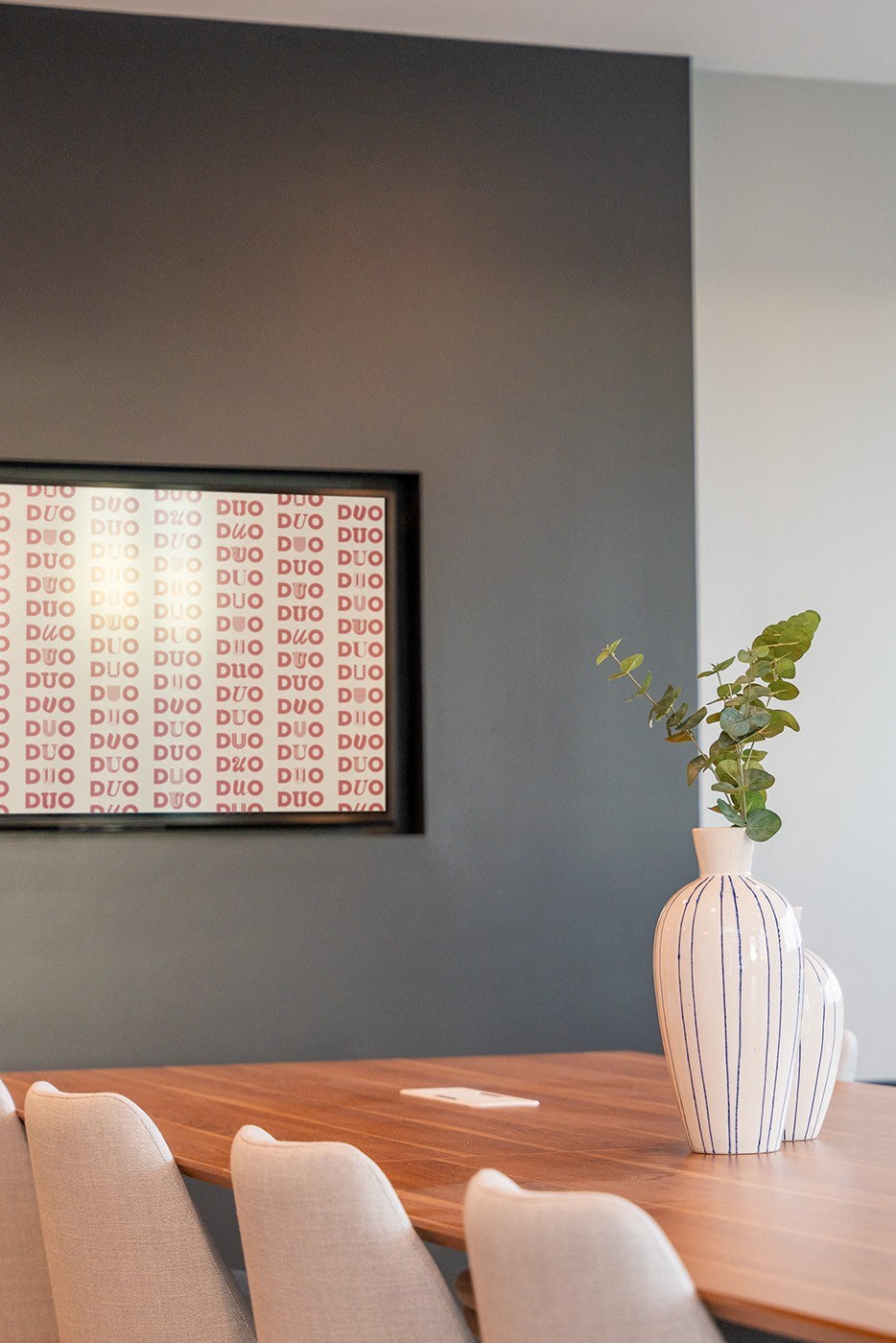 A white vase with a green plant sits on a wooden table in front of a wall with a framed picture.