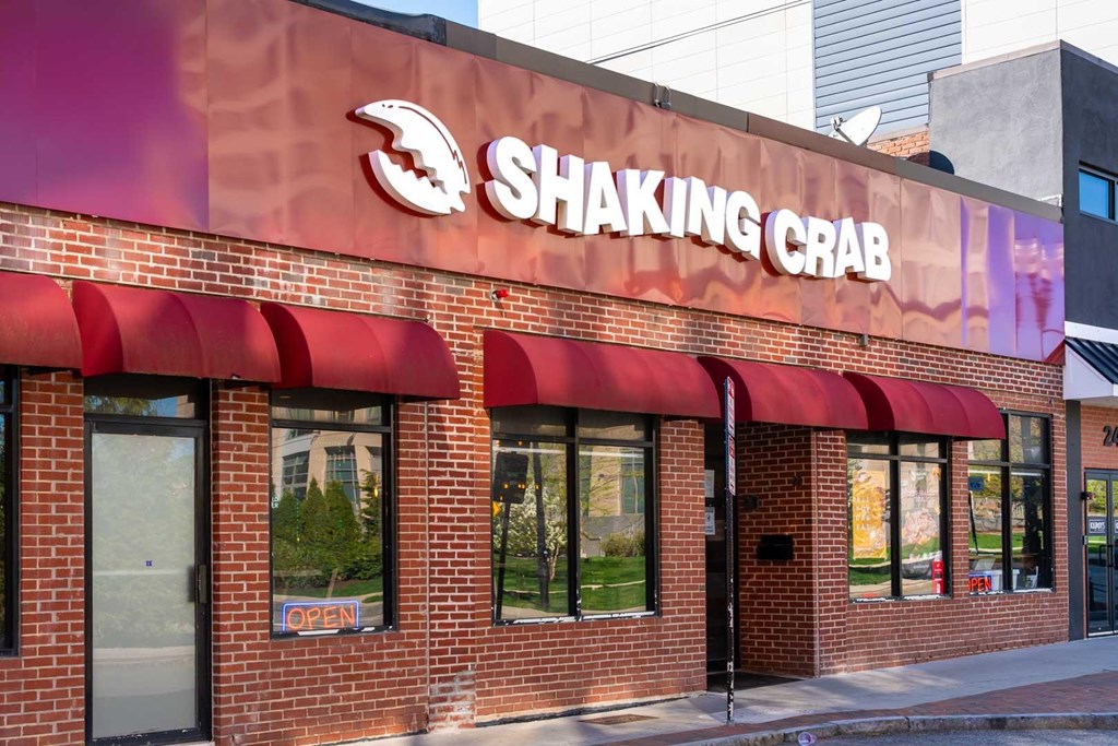 The exterior of a Shaking Crab restaurant with a red awning and a blue sign on the window.
