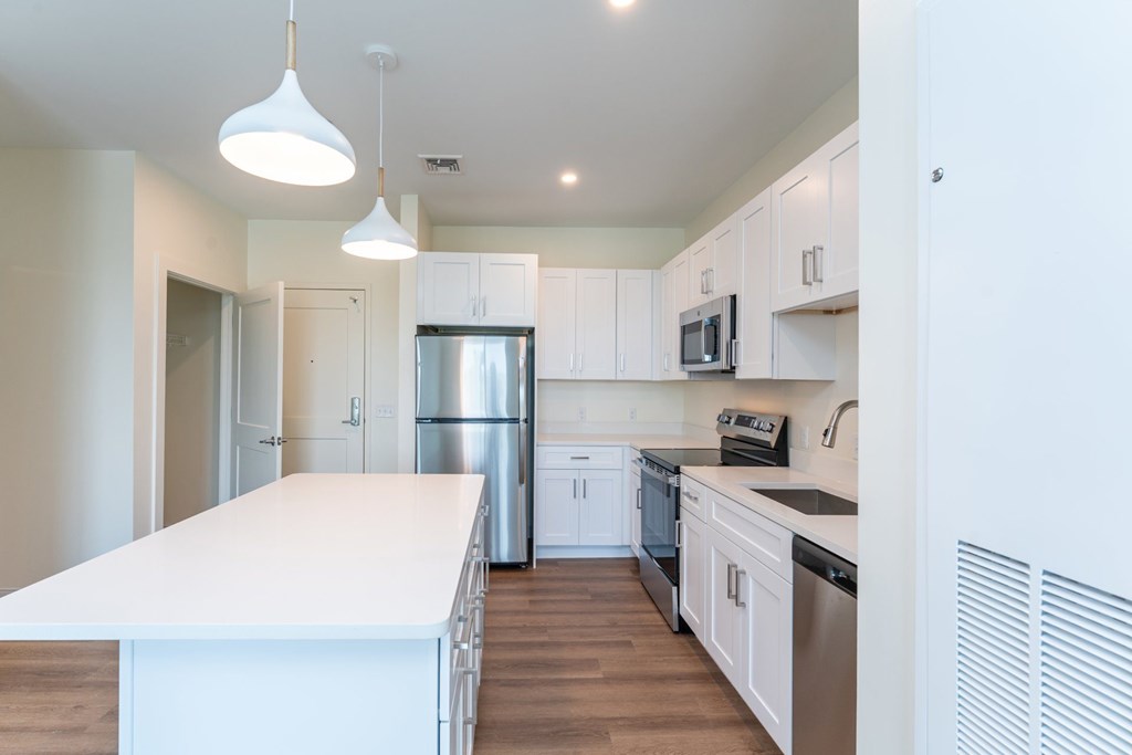 Bright, modern kitchen with a long white island, stainless-steel appliances and pendant lighting beneath white Shaker cabinets.