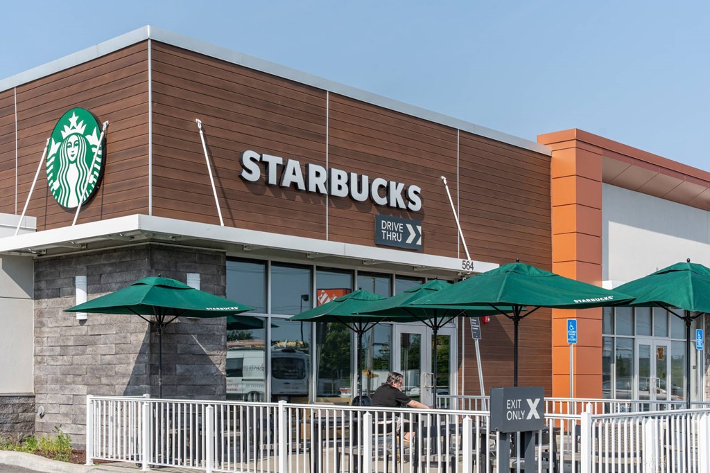 Starbucks café with wood-accent façade, drive-thru sign and patio tables shaded by green umbrellas.