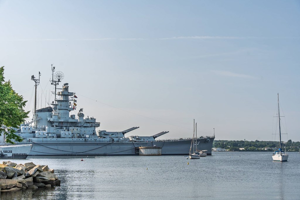Historic battleship anchored on a calm river alongside small sailboats and tree-lined shoreline.