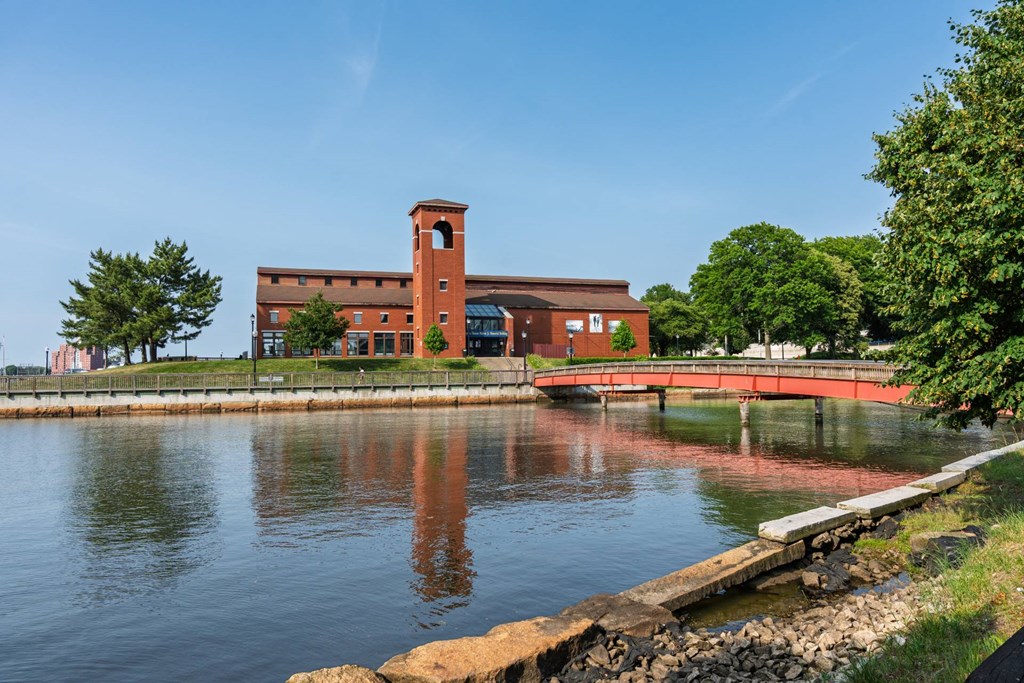 Brick waterfront museum with tower reflected in the river, connected by a red pedestrian bridge beneath a clear sky.