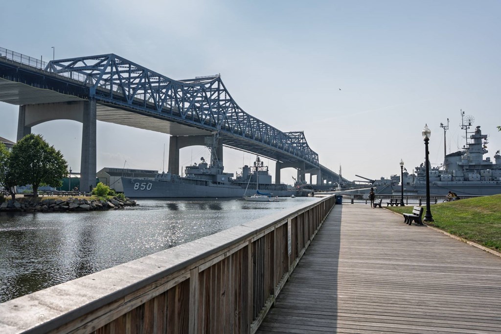 Boardwalk beneath a steel truss bridge, looking toward moored naval ships along the sparkling river.