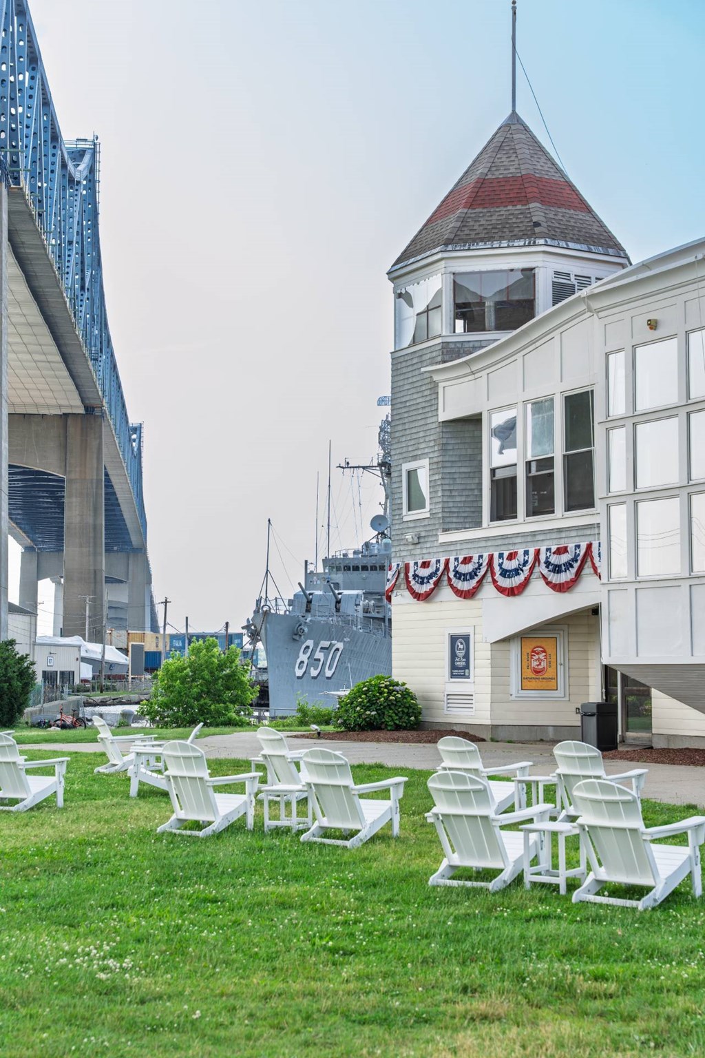 Lawn with white Adirondack chairs beside a carousel pavilion draped in patriotic bunting, with destroyer 850 and bridge backdrop at Battleship Cove