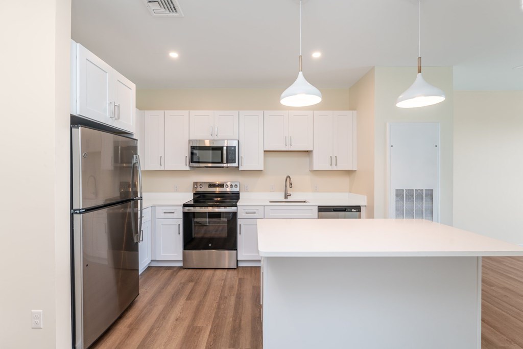 Open kitchen lined with white Shaker cabinets, quartz island workspace and twin pendant lights, plus stainless fridge and range.