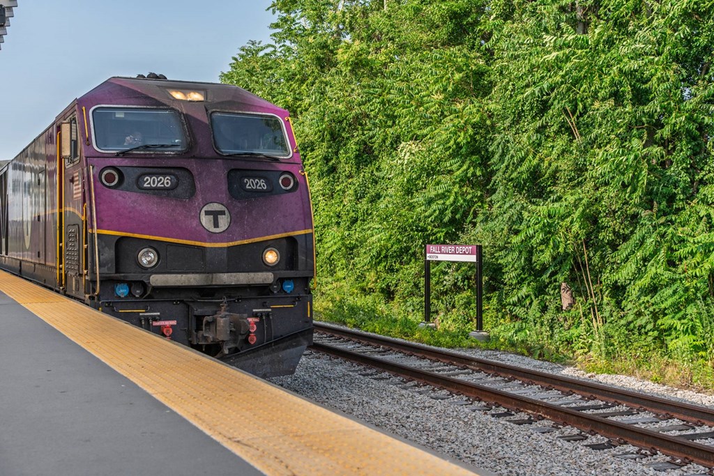 Purple MBTA commuter-rail locomotive arriving at Fall River Depot, framed by tree-lined tracks and platform edge.