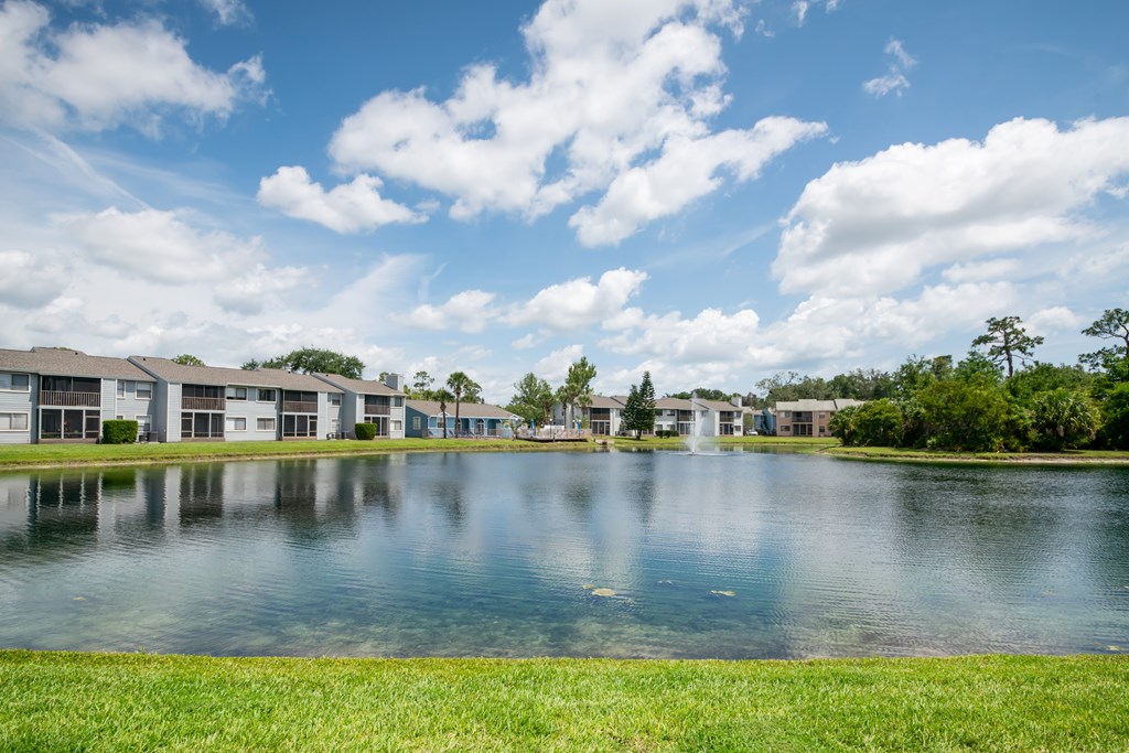 A serene lake in front of a row of houses under a blue sky with clouds Fishermans Landing Apartments in Ormond Beach, FL.