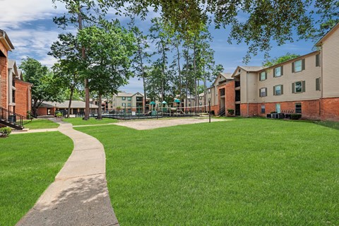A grassy area with a concrete walkway and apartment buildings in the background.