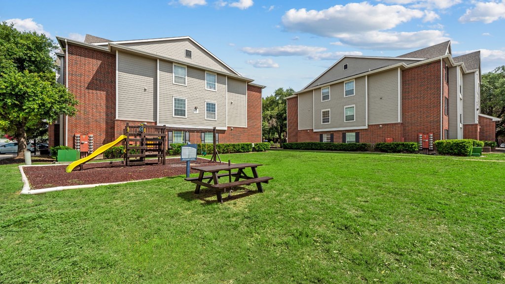 A grassy area in front of apartment buildings with a picnic table and playground.
