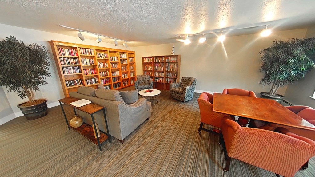 A room with a long bookshelf, a couch, a chair, a table, and a potted plant.