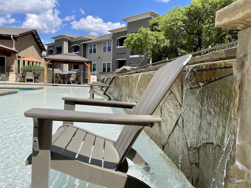 a lounge chair sits on the edge of a pool with a waterfall and buildings in the background