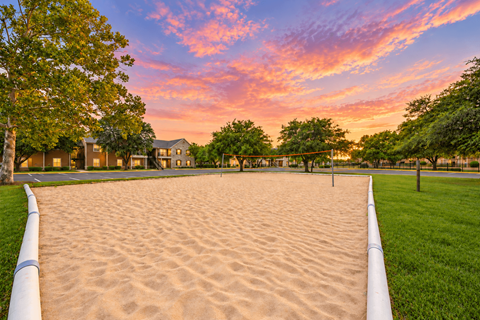 A sandy area with a white fence and green grass in the foreground and houses in the background.