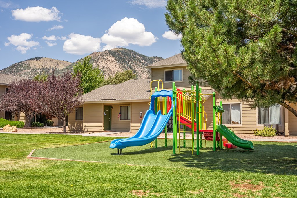 A playground with a blue slide and a green tree in front of a house.