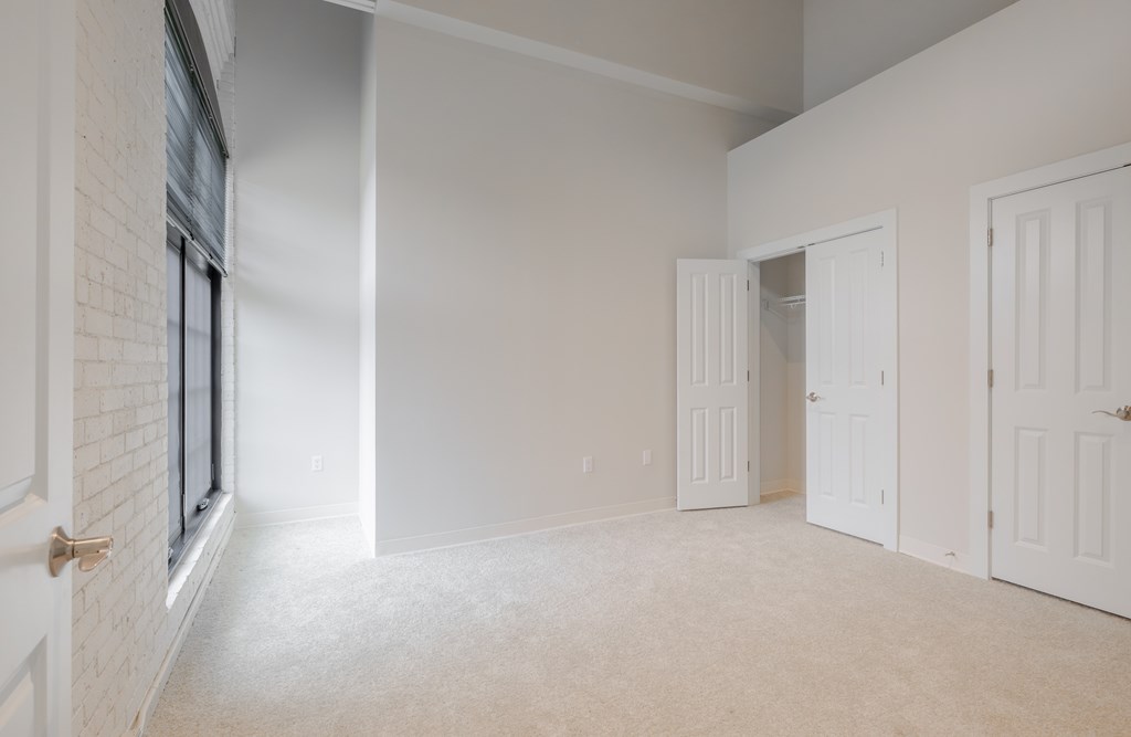 A room with white walls and carpeted floor with a window on the left and three doors on the right.