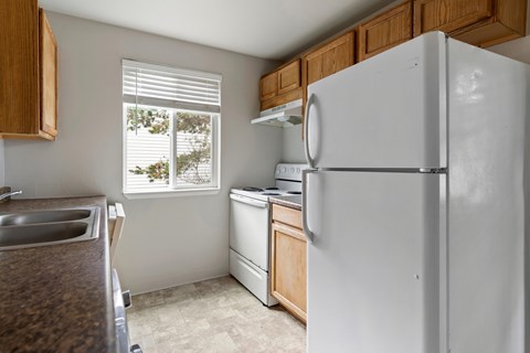 A white refrigerator stands in a kitchen with wooden cabinets and a window with blinds.