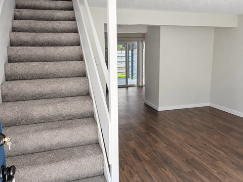 A staircase with a grey carpeted runner and white handrail leads to a bright room with a wooden floor and a glass door.