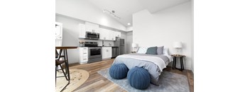 A modern bedroom with a bed, two blue knitted poufs, and a kitchen area with white cabinets and appliances.