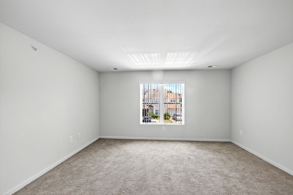 A large empty room with a carpeted floor and a window showing a cityscape outside.
