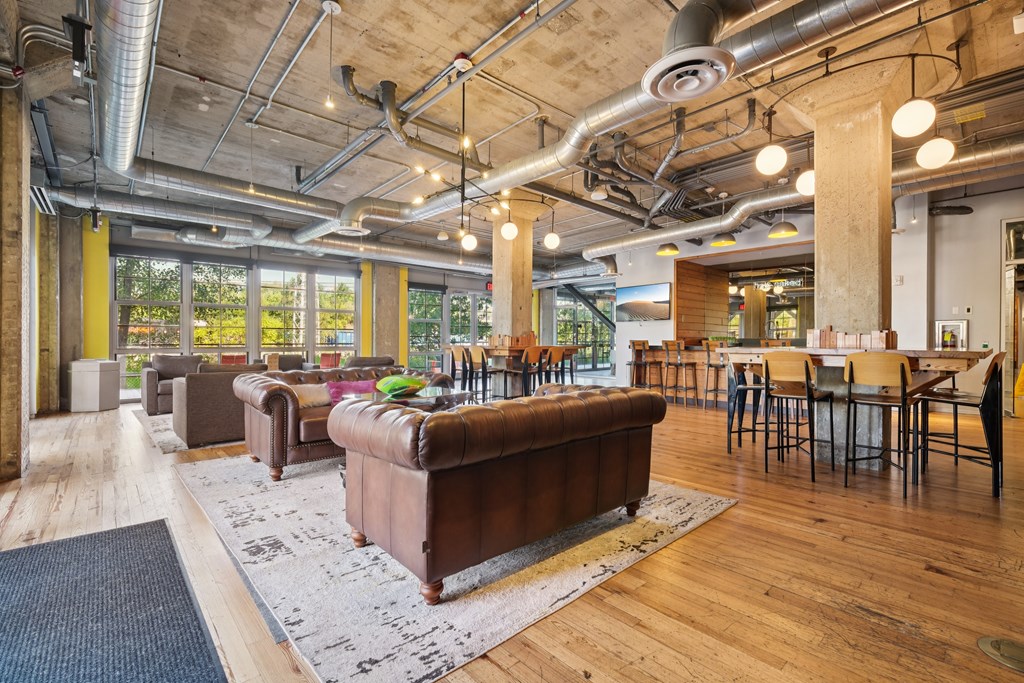 A large brown leather couch sits in the middle of a room with wooden floors and a ceiling full of pipes.