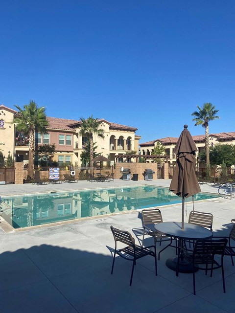 A table with chairs and an umbrella is in the foreground of a pool area.