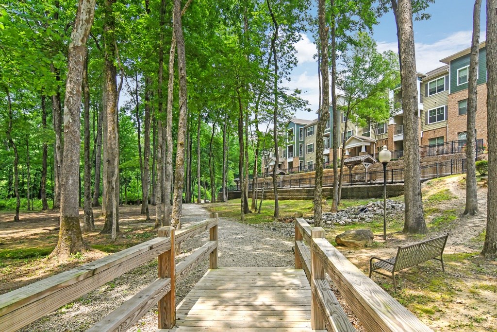 a pathway through the woods with a bench and a building in the background at Faulkner Flats Apartment Homes, Oxford, MS