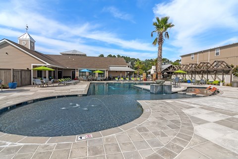 A large outdoor swimming pool surrounded by a tiled patio and a building with a clock tower in the background.