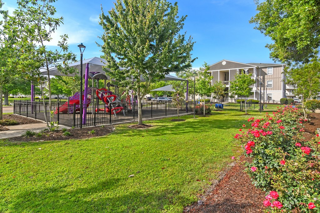A playground with a red slide and a purple canopy is surrounded by a green lawn and a fence at Gulf Hills Apartment Homes in MS 39564