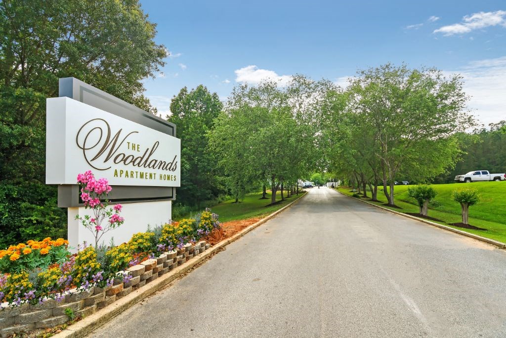 a street with a sign that says the woodlands with trees and flowers in front of it at The Woodlands Apartment Homes, Meridian, MS, 39301