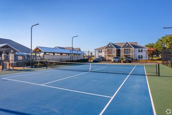 A tennis court with a blue surface and white lines, surrounded by a fence and a building in the background at Reserve at Park Place Apartment Homes in Bossier City, LA