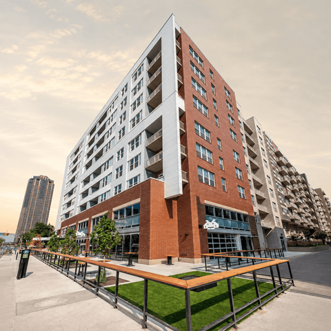 A modern multi-story building with a red brick facade and a balcony with a wooden railing.