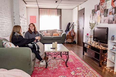 two women sit in a living room with a red rug and a television at University Commons Apartments, Oakland, Pittsburgh