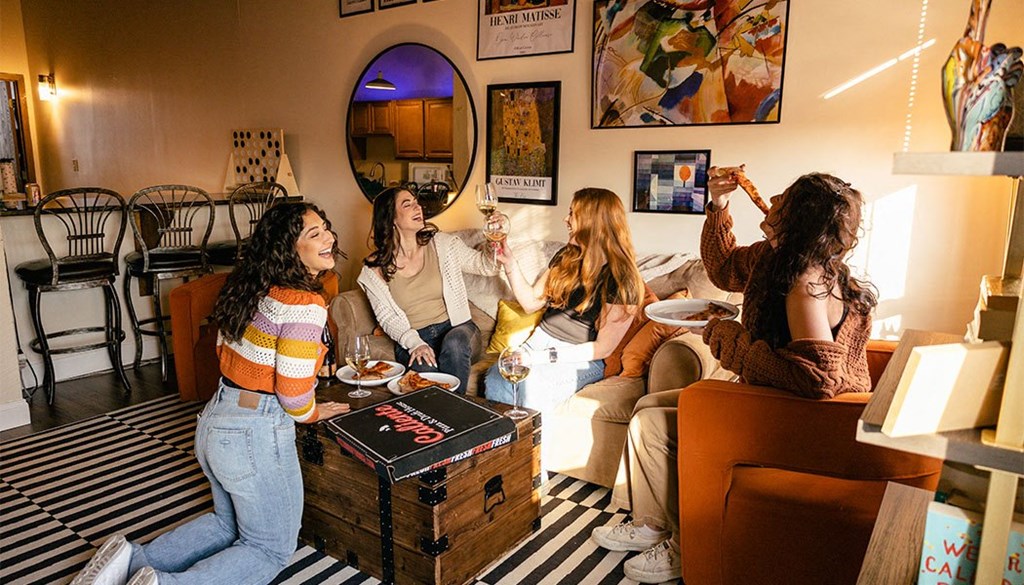 a group of women sitting in a living room drinking wine and eating food