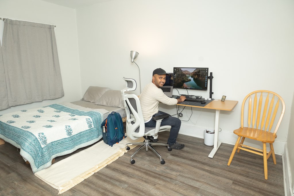 A man is sitting at a desk in a room with a bed and a chair.