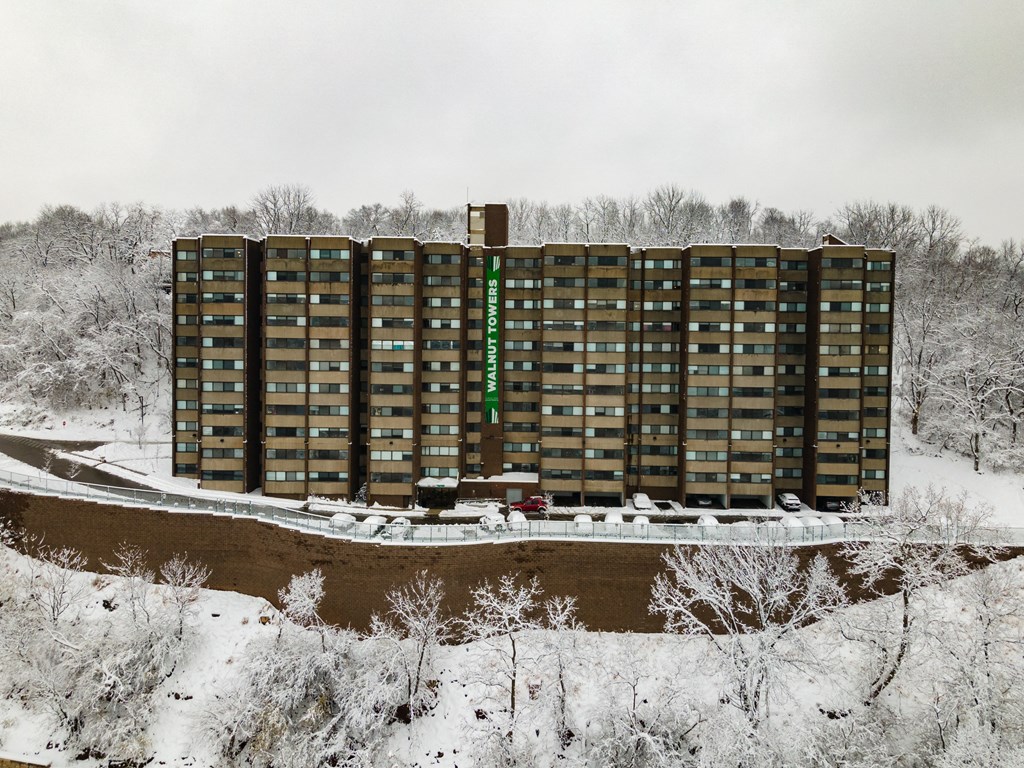 A tall building with a green sign on the side stands in front of a snowy landscape at Walnut Towers at Frick Park, Pittsburgh, PA