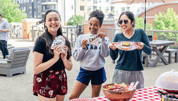 Three people holding food items in front of a picnic table.