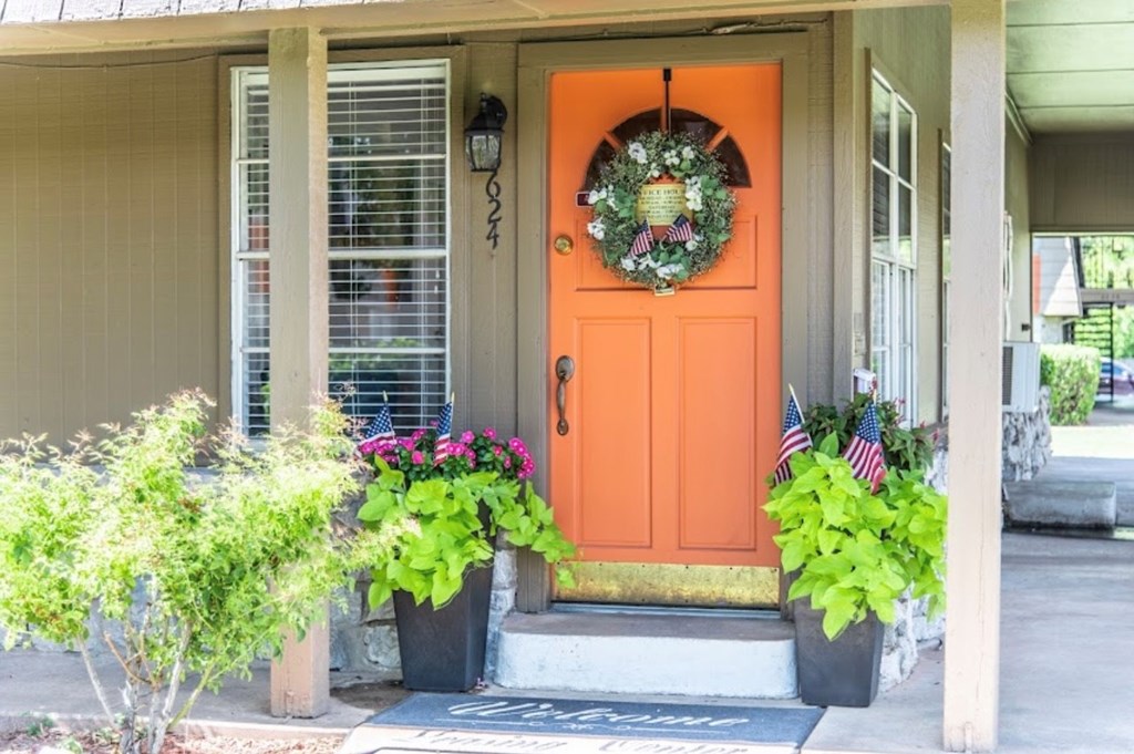 A bright orange door with a wreath and two potted plants on either side.