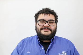A man with glasses and a beard wearing a blue shirt with the name Alex on it.